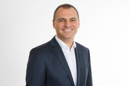 Portrait von Dirk Simons. Er trägt ein weißes Hemd sowie einen dunkelblauen Blazer und steht lächelnd vor einer weißen Wand.