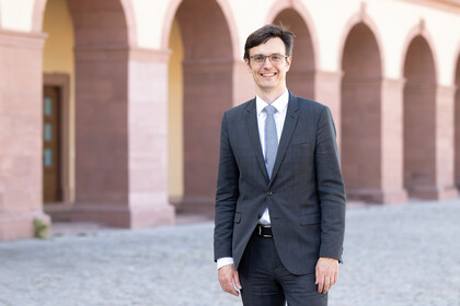 Eine Person trägt ein weißes Hemd, einen grauen Blazer sowie einen grau gemusterten Blazer und steht vor dem Schloss. Die Person heißt Erik Röder.
