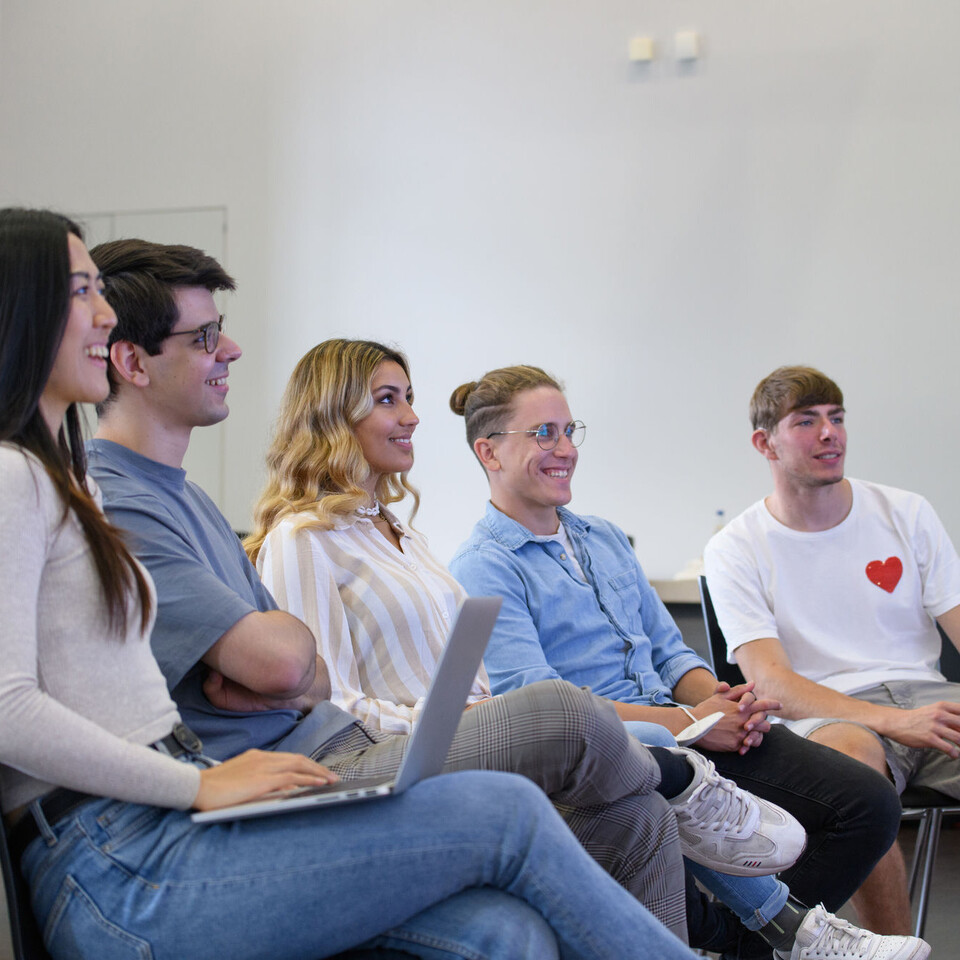 Studierende sitzen auf Stühlen und schauen nach vorne an die Tafel.