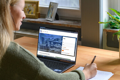 Studierende mit langen blonden Haaren sitzt vor einem Laptop und macht sich daneben Notizen auf einem Block 