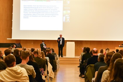 Blick aus dem Mittelganz zwischen dem Publikum auf Prof. Dr. Dr. h.c. Dieter Birnbacher, der auf eine Stelle links im Publikum schaut und gestikuliert. Prof. Birnbacher steht auf der leicht erhöhten Bühne links des Rednerpults, ist bekleidet mit dunkelblauen Anzug und hellblauem Hemd, trägt eine Brille und hat ein Funkmikrofon umgehängt. Etwas links versetzt davon sitzt in schwarzem Anzug, weißem Hemd und in ebenfalls diesen Farben gestreifter Krawatte bei umgehängten Funkmikrofon Prof. Dr. Bernward Gesang. Den Hintergrund des Fotos bildet eine dunkle Holzvertäfelung und helle Projektionsfläche mit angeschnittener projizierter Powerpoint-Folie. 