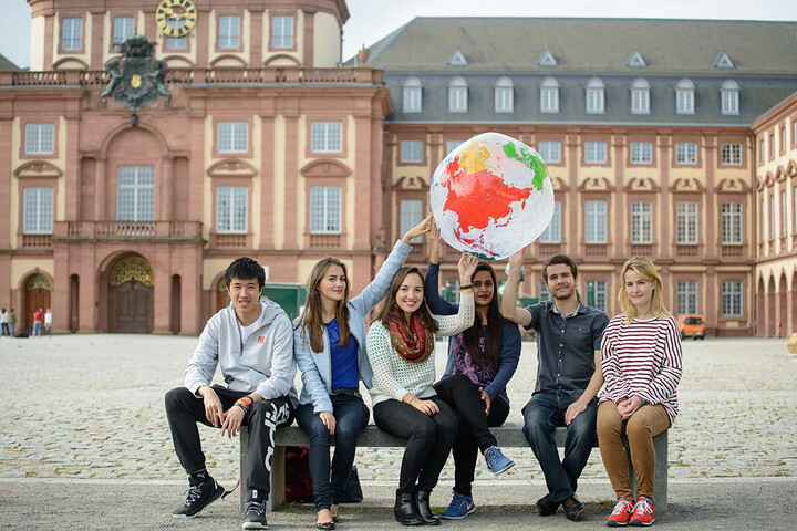 Six students sit on a bench in the courtyard of honor in front of Mannheim Palace and hold a globe balloon above their heads. 