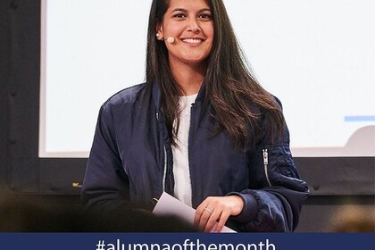 Sukhmani Nagi hat lange braune Haare und hat ein Mikrofon aSie trägt ein weißes Oberteil und eine dunkelblaue Jacke. Darunter der Hashtag Alumna of the month.