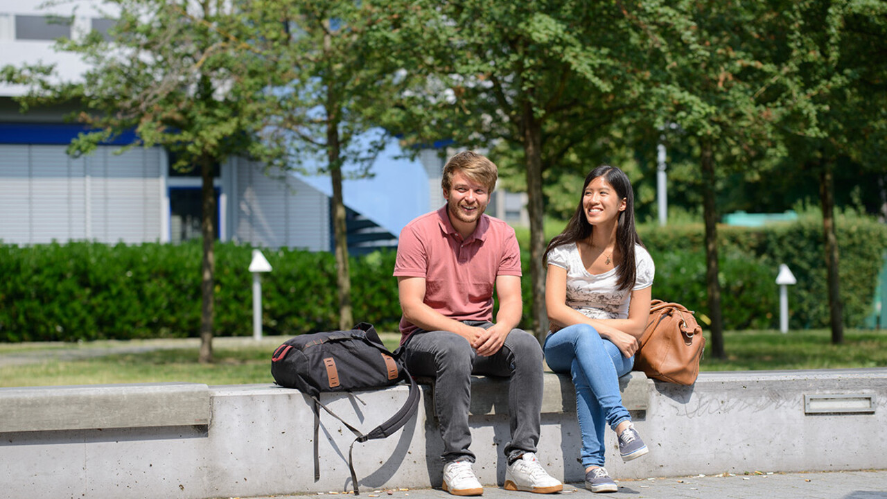 Zwei Studierende sitzen nebeneinander auf einer Bank vor dem A5-Gebäude.