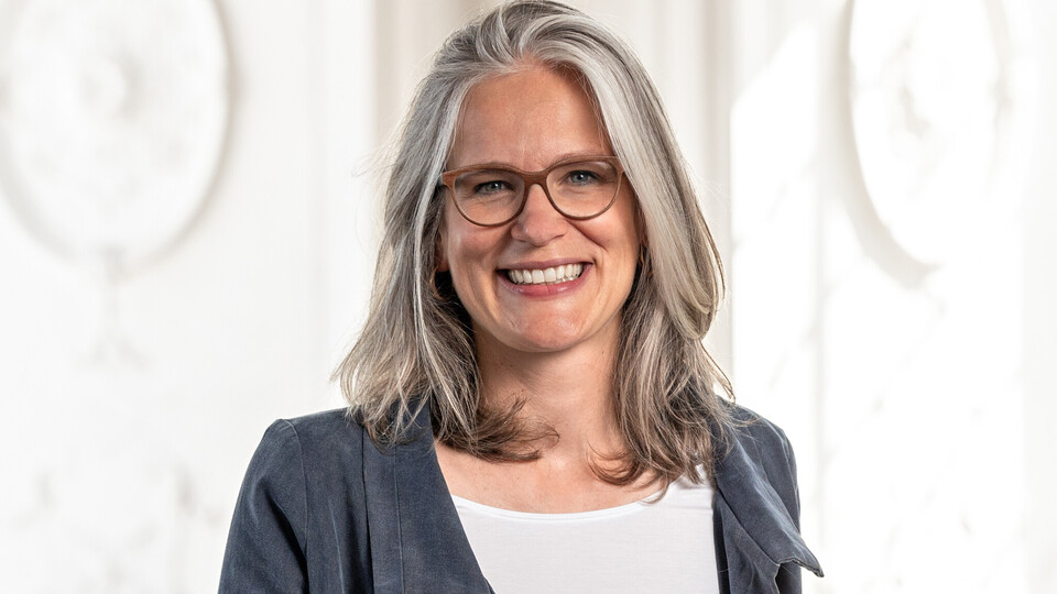 Portrait von Jutta Mata. Sie trägt ein weißes Oberteil mit einem grauen Blazer und lächelt. 
