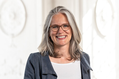 Portrait von Jutta Mata. Sie trägt ein weißes Oberteil mit einem grauen Blazer und lächelt. 