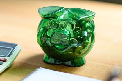 Grünes Plastiksparschwein auf einem Tisch. Davor ein Schreibblock und ein Taschenrechner.