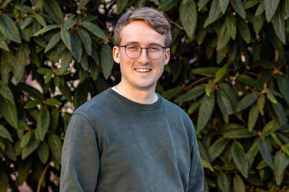 Gerrit von Zedlitz-Neukirch steht lächelnd mit Brille und grünem Pullover vor einem Hintergrund aus grünen Blättern.