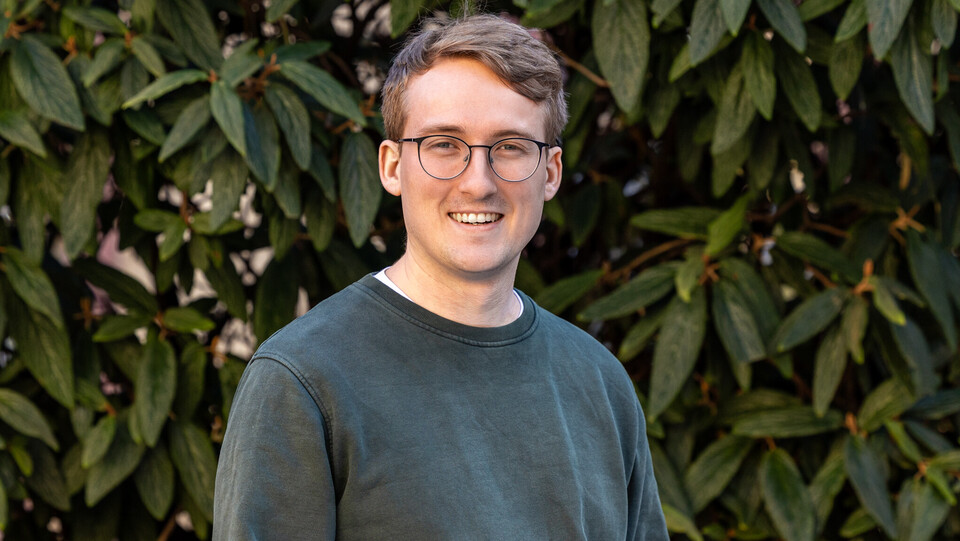 Gerrit von Zedlitz-Neukirch steht lächelnd mit Brille und grünem Pullover vor einem Hintergrund aus grünen Blättern.