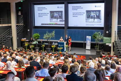 Das Bild zeigt ein Bild von der Absolvierendefeier im Hörsaal A. Auf der Bühne stehen drei Personen, die die Abschlussrede halten, im Publikum sitzen Absolvierende und ihre Begleitungen. Das Publikum ist von hinten zu sehen.