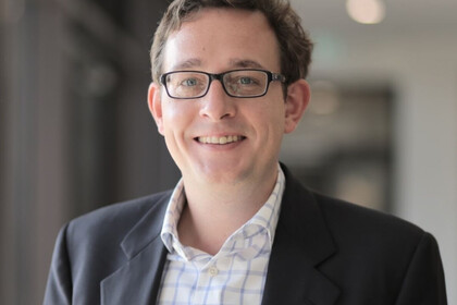 Porträt des "Alumnus of the month" Prof. Dr. Philipp Sandner. Er hat kurze braune Haare und lächelt in die Kamera. Er trägt ein schwarzes Sakko, ein weißes Hemd mit blauem Karomuster und eine Brille. Die Brille hat ein dunkles eckiges Gestell. Link: Instagram-Post Alumnus of the month
