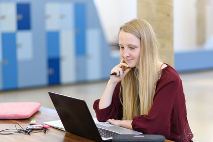 Eine Studentin sitzt mit ihrem Laptop an einem Tisch und betrachtet etwas auf dem Bilschirm.