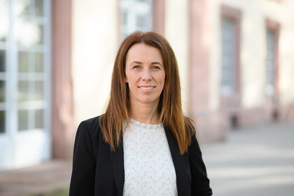 Alexandra Niessen-Ruenzi trägt eine weiße Bluse mit einem schwarzen Blazer und steht vor einem Gebäudeteil der Universität.