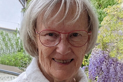 Beate Kohler hat graue Haare und eine rot umrandete Brille. Im Hintergrund sieht man lilanen Flieder. 