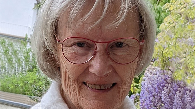 Beate Kohler hat graue Haare und eine rot umrandete Brille. Im Hintergrund sieht man lilanen Flieder. 
