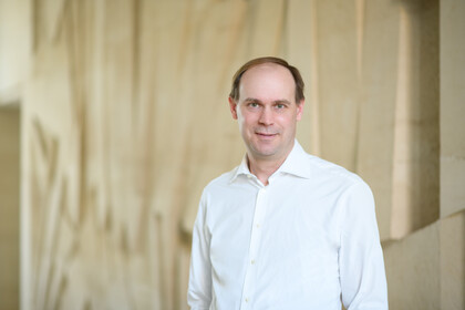 Portrait von Jannis Bischof. Er trägt ein weißes Hemd und steht vor einer beigen Wand.