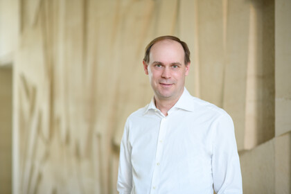 Portrait von Jannis Bischof. Er trägt ein weißes Hemd und steht vor einer beigen Wand.
