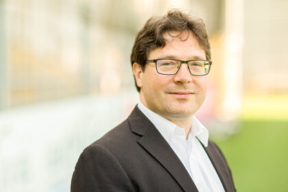 Portrait von Philipp S. Fischinger. Er trägt ein weißes Hemd sowie einen braunen Blazer und steht auf einer Rasenfläche.