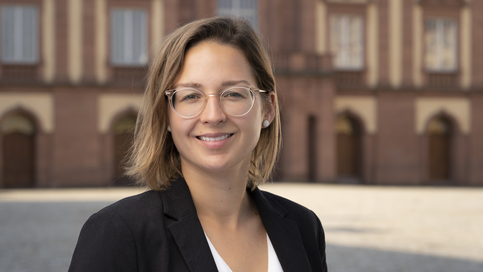 Isabella Heppting hat schulterlange blonde Haare und eine Brille. Sie trägt eine weiße Bluse und einen dunklen Blazer. Im Hintergrund ist das Schloss zu sehen.