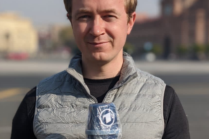 Demian von Osten hat kurze blonde Haare. Er trägt ein schwarzes Oberteil mit einer grauen Weste darüber und hält ein blaues Mikrofon in der Hand. Darunter steht "Alumus of the month".
