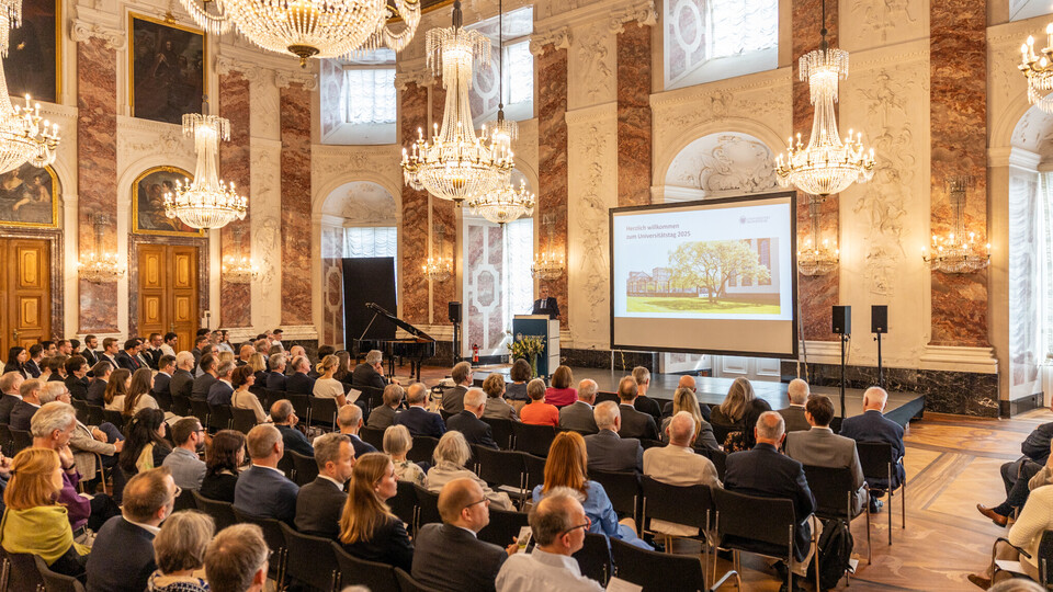 Blick in den Rittersaal. An der Decke hängen Kronleuchter, das Publikum sitzt in Stuhlreihen vor der Bühne und ist von hinten zu sehen. Auf der Bühne steht Professor Fetzer, neben ihm ist eine Leinwand zu sehen.