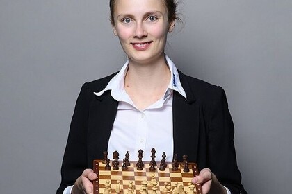 Portrait von Student*in Josefine Heinemann (blonde Haare, weißes Hemd, schwarzes Jackett), ein Schachbrett vor sich haltend.
