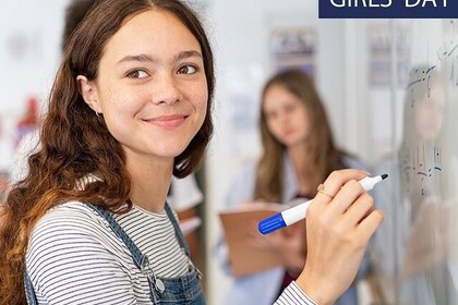 Ein Mädchen mit geringeltem Oberteil und Latzhose schreibt mit einem Filzstift eine mathematische Formel auf eine weiße Tafel. Link: Insta Girls' Day