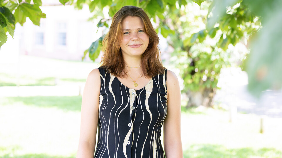 Clara Schünemann. Sie steht unter einem Baum und lächelt, dabei trägt sie ein schwarz-beige gemustertes Oberteil.