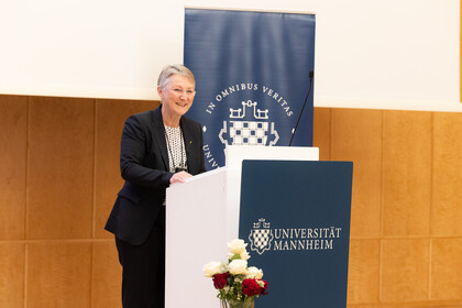Portrait von Barbara Windscheid. Sie trägt eine weiße Bluse und einen schwarzen Blazer und steht an einem Rednerpult.