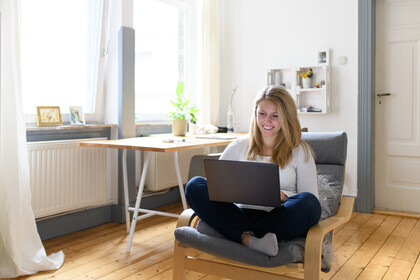 Frau mit langen blonden Haaren in blauen Jeans und weißem Pulli im Schneidersitz auf einem Sessel schaut lächelnd auf ein Laptop auf Ihren Beinen. Den Hintergrund bildet ein helles Zimmer mit Holzfußboden und Fenstern auf der linken Bildseite.