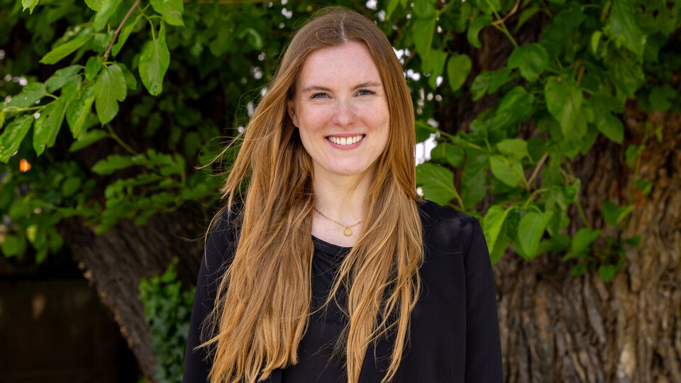 Verena Weller trägt ein dunkles Jacket und lächelt sympathisch in die Kamera. Im Hintergrund sind grüne Blätter.