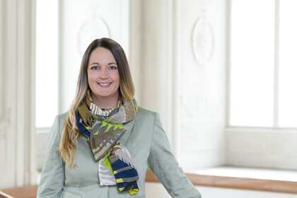 Portrait von Laura Marie Edinger-Schons. Sie trägt einen bunten Schal sowie einen zart grünen Blazer und steht vor einem Fenster.