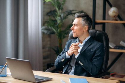 Ein Mann in einem Anzug sitzt an einem Schreibtisch. Auf dem Tisch steht ein Laptop. Der Mann schaut aus dem Fenster und faltet nachdenklich die Hände.