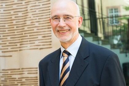 Thomas Puhl hat kurze graue Haare und eine Brille. Er trägt einen schwarzen Anzug mit einer schwarz-gelb-gestreiften Krawatte.