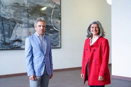 Daniel Roggenkamp ht kurze graue Haare und trägt einen hellblauen Anzug. Caroline Mary hat graue mittellange Haareund trägt einen roten Mantel. Sie stehen nebeneinander und blicken in die Kamera.