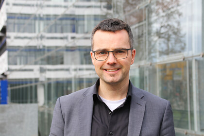Porträt von Prof. Debus. Er steht vor dem Gebäude A5 und trägt kurze Haare, eine Brille und ein graues Sacko.