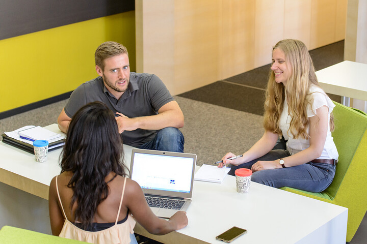 Zwei Studentinnen und ein Student sitzen gemeinsam mit Lernmaterialien an einem Tisch und unterhalten sich.