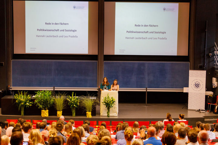 Two women stand on stage and give a speech. Both are standing at the lectern, the guests are sitting in the rows of the lecture hall in front of them.