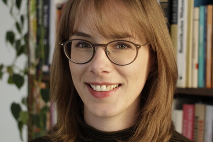 Portrait von Almuth Ebke. Sie trägt einen schwarz rot gestreiften Pullover und steht lächelnd vor einem Bücherregal.