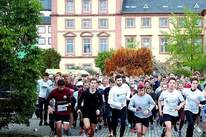 Eine Menschengruppe in Sportkleidung absolviert einen Lauf vor der Mensa. Die Teilnehmer*innen laufen auf die Kamera zu, im Hintergrund sind Universitätsgebäude und die Jesuitenkirche.