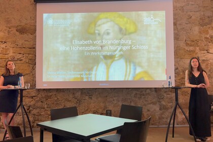 Zwei Frauen stehen an zwei Stehtischen neben einer Leinwand mit der Präsentation 'Elisabeth von Brandenburg – eine Hohenzollern im Nürtinger Schloss' in einem gemauerten Kellergewölbe