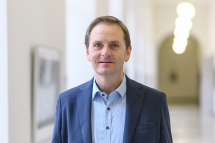 Portrait von Oliver Dickhaeuser. Er trägt ein hellblaues Hemd sowie einen blauen Blazer und steht in einem Flur der Uni.