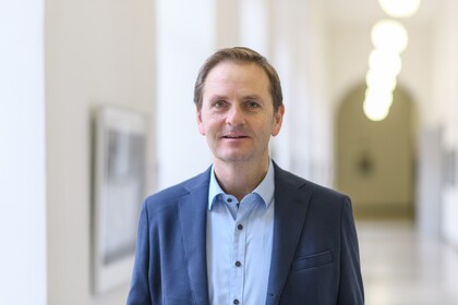 Portrait von Oliver Dickhaeuser. Er trägt ein hellblaues Hemd sowie einen blauen Blazer und steht in einem Flur der Uni.