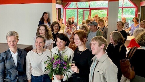 Bild einer Gruppe die alle lachend in die Kamera schauen. In der Mitte eine Frau mit einem Blumenstrauß.