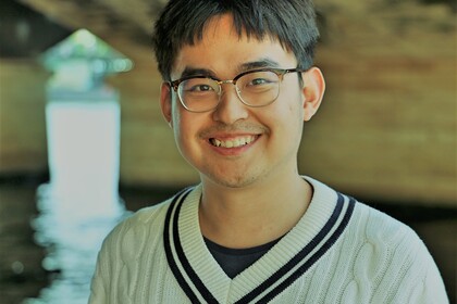 Youzhe Heng hat kurze schwarze Haare. Er lächelt und trägt eine Brille und einen weißen Strickpullover mit V-Ausschnitt.