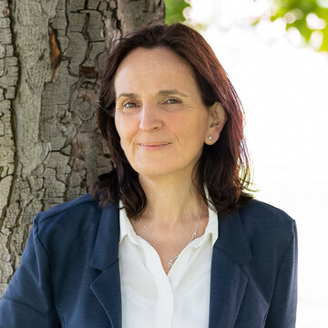 Frau mit schulterlangen braunen Haaren, weißer Bluse, dunkelblauen Blazer und dunkelblauer Hose steht auf der Mensawiese unter einem Baum und lächelt.