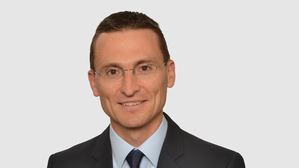 Prof. Brauer hat kurze Haare und eine Brille. Er trägt ein dunkles Jacket, ein hellblaues Hemd und eine dunkelblaue Krawatte.