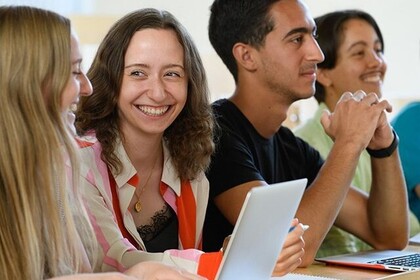 Vier Studierende sitzen im Hörsaal und lächeln. Vor ihnen liegen Laptops und Schreibutensilien.