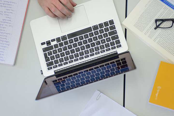 Man blickt von oben auf eine Hand die einen Laptop bedient. Um den Laptop herum liegen Papiere, Bücher und ein Notizblock verteilt.