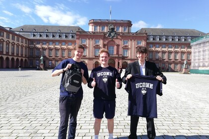 Drei Studierende stehen auf dem Ehrenhof vor dem Schloss und halten T-Shirts und Pullis der UConn Business School in die Kamera.